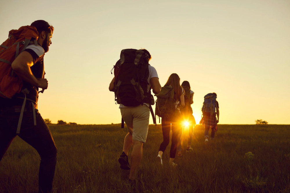 Pessoas andando no campo com mochilas, com o sol nascendo no horizonte